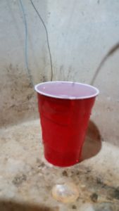 Cup with two wires hooked up, water running into the top, and water overflowing down the side.  Cup is sitting in a laundry room slop sink.